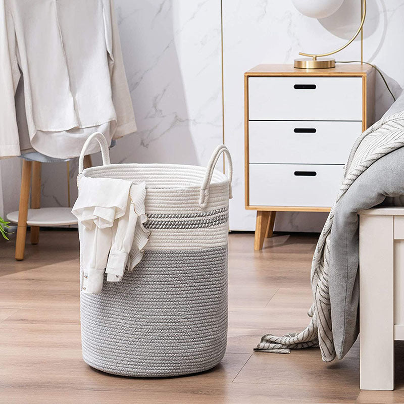 Grey and white two-tone cotton rope laundry basket used as a bedroom organizer next to a modern nightstand and bed. Handcrafted decorative storage bin for blankets, toys, and nursery essentials.