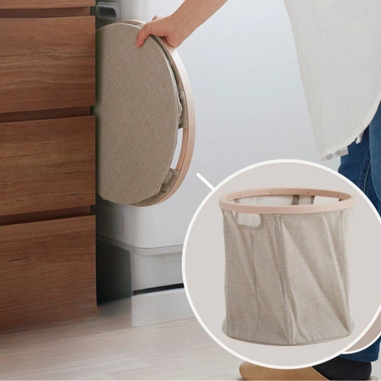 Person collapsing a wooden ring laundry basket flat for storage next to a dresser, with inset showing the expanded basket.