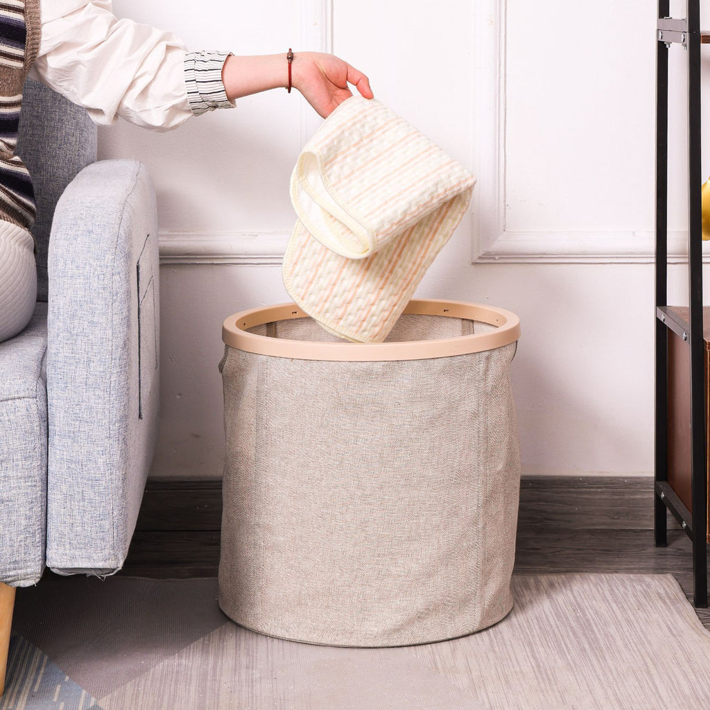 Person dropping a knit sweater into a wooden ring canvas laundry basket placed beside a sofa in a living room.