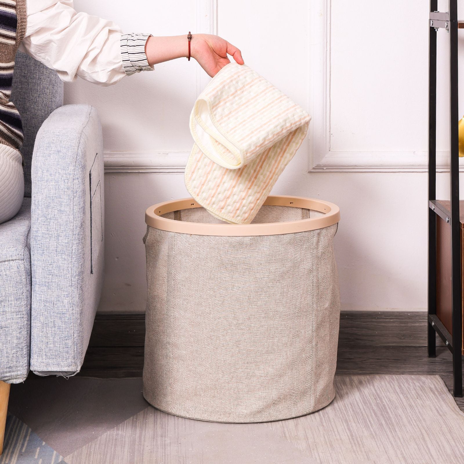 Person dropping a knit sweater into a wooden ring canvas laundry basket placed beside a sofa in a living room.