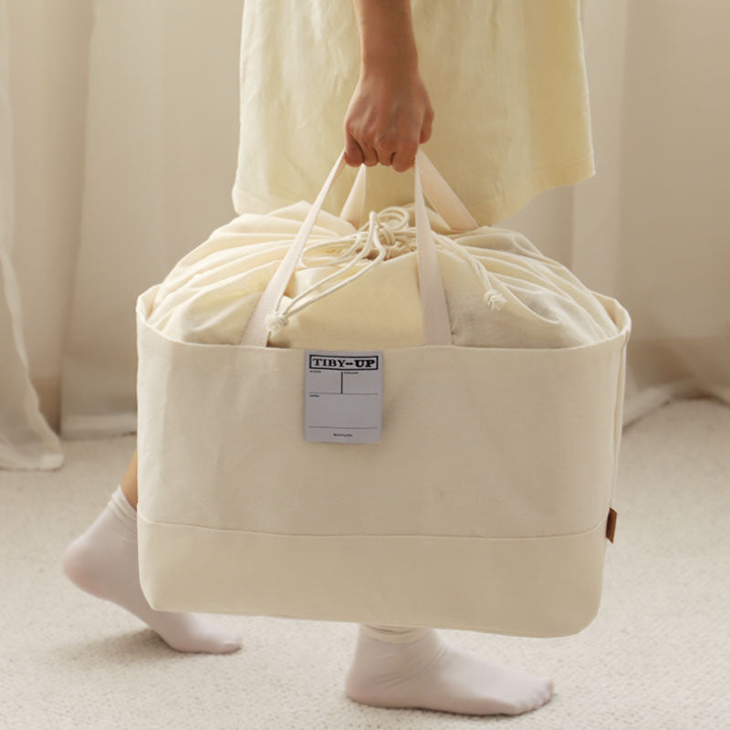 Person carrying a large cream canvas drawstring storage bag filled with bedding by its sturdy handles.