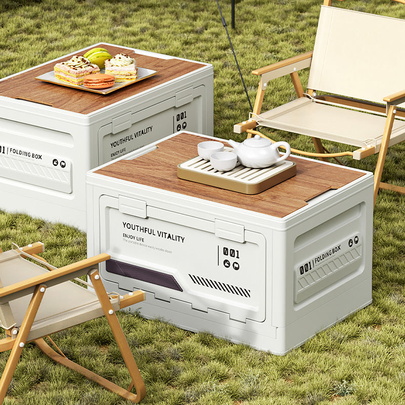 Two white foldable storage boxes with wooden tops used as outdoor tables on a grass lawn. One holds a white tea set, the other displays macarons and pastries. Beige folding chairs complete the cozy picnic setup