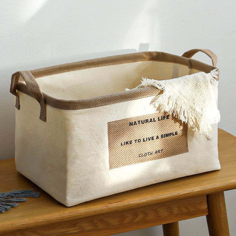 Foldable cotton-linen storage basket with burlap handles and fringe throw, placed on a wooden table in natural light.