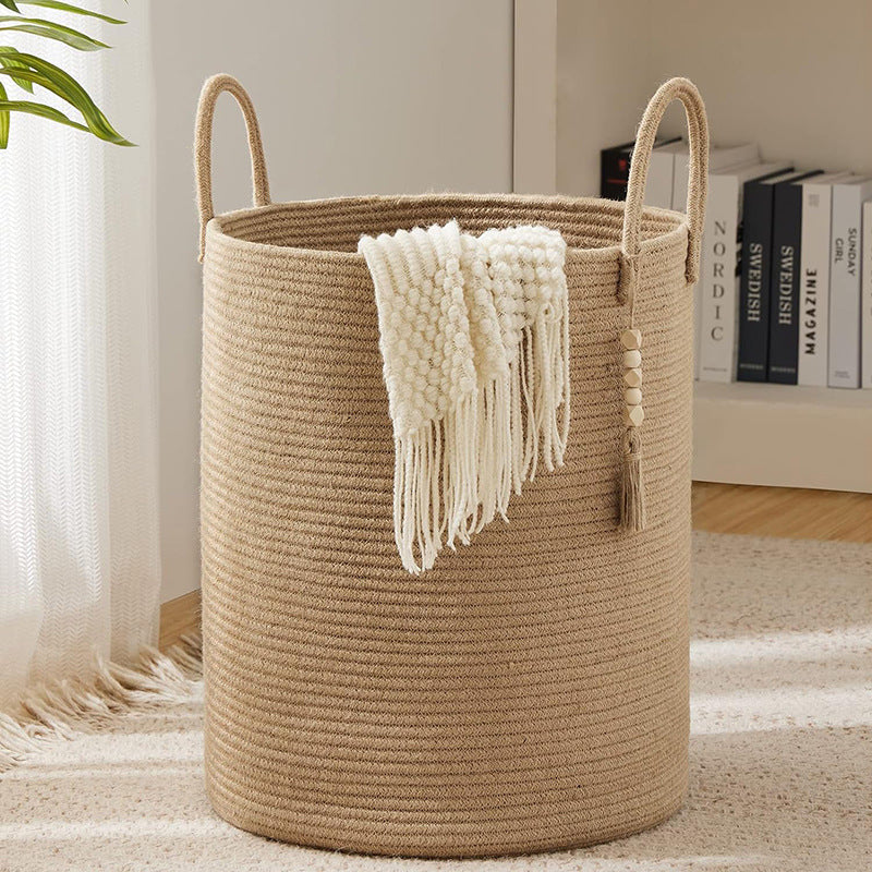 Large khaki woven cotton rope storage basket with dual loop handles and boho tassel accent, placed on a white carpet in a bright living room with a white knit blanket draped over the side.