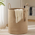 Large khaki woven cotton rope storage basket with dual loop handles and boho tassel accent, placed on a white carpet in a bright living room with a white knit blanket draped over the side.