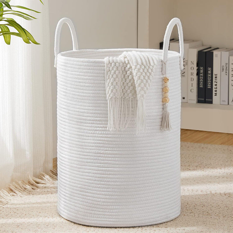 Large white woven cotton rope storage basket with loop handles and boho wood bead tassel accent, with a white knit throw draped over the side, styled in a bright living room.