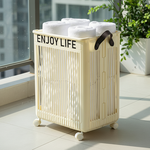 Cream white foldable rolling storage hamper filled with rolled white towels, placed on a sunny balcony tile floor beside a large potted green plant with city views in the background