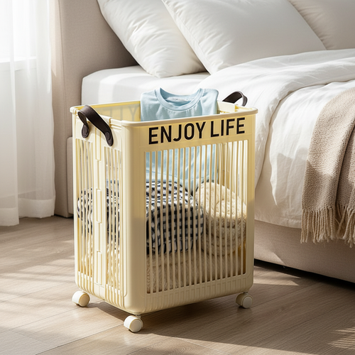 Cream white foldable rolling laundry hamper with leather handles and "Enjoy Life" slogan, filled with clothes, placed on a wood floor beside a white bed in a bright bedroom