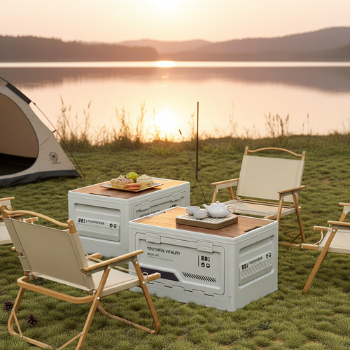 Two white foldable storage boxes (YOUTHFUL VITALITY) with wooden board tops used as outdoor tables, set on a grassy lakeside at golden sunset. One box holds a white tea set on a wooden tray, the other displays pastries and macarons. Beige folding wooden chairs surround the setup, with a dome camping tent visible on the left and a serene lake and mountain backdrop glowing in the warm evening light.