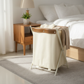 Cream canvas laundry hamper with tan leather trim and X-frame stand, placed beside a bed in a bright, minimalist bedroom.