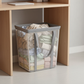 Mesh laundry hamper with gray handles stored under a wooden shelf, filled with light clothing.