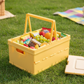 Yellow foldable storage crate with handle, filled with fresh fruits and snacks, on green grass outdoors.